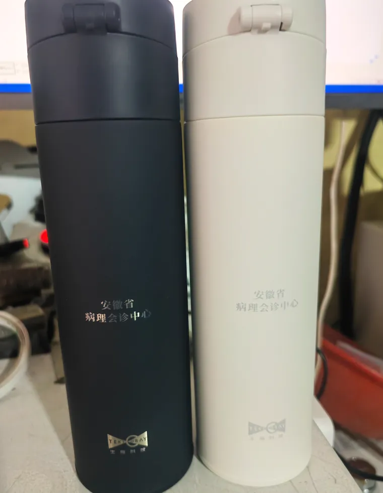 合肥激光打标代工实拍 · 黑色与米白色不锈钢保温杯曲面激光打标「安徽省病理会诊中心」，深色杯打浅字、浅色杯打深字，弧面不变形不起皮
